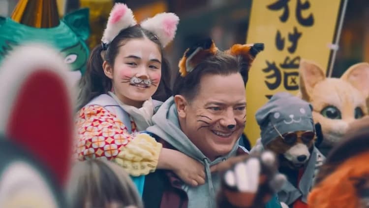 Phillip is giving Mia a piggyback ride. They are in the middle of a large crowd. Both have fox ears and face paint on. Numerous people in the background are wearing fox masks. A yellow banner is in the background.