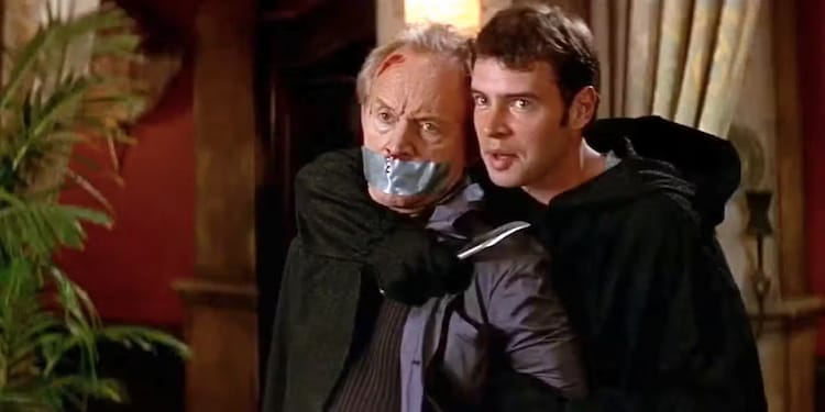 Roman Bridger, right, is holding a knife to Milton’s throat. Roman is wearing a Ghostface outfit but is not wearing a mask.